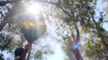 Kişi tarafından tutulan LGBTQ + gurur bayrağı renginde kalp şeklinde bir balon. Güneş ışınları eşitliği, cinsel özgürlüğü, cinsiyet saygısını ve dahil edilmeyi simgeliyor.