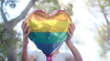 Kişi tarafından tutulan LGBTQ + gurur bayrağı renginde kalp şeklinde bir balon. Güneş ışınları eşitliği, cinsel özgürlüğü, cinsiyet saygısını ve dahil edilmeyi simgeliyor.