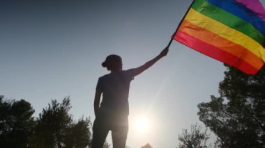 Gün batımında dalgalanan LGBTQ + bayrağıyla yalnız genç bir kadın. Eşitlik, eşitlik, dahil etme, LGBTQ konsepti. Aşk aşktır, ayrımcılık yok, özgürlüğe evet