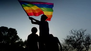 Anne ve iki çocuk gün batımında bir dağın tepesinde LGBTQ + bayrağı sallıyorlar. Aile, çeşitlilik, kapsama ve eşitlik kavramı. Gurur, lezbiyen anne ve çocuklar. Aşk aşktır.