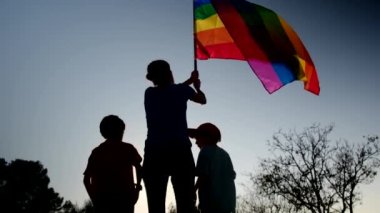 Anne ve iki çocuk gün batımında bir dağın tepesinde LGBTQ + bayrağı sallıyorlar. Aile, çeşitlilik, kapsama ve eşitlik kavramı. Gurur, lezbiyen anne ve çocuklar. Aşk aşktır.