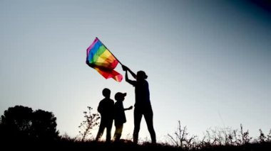 Anne ve iki çocuk gün batımında bir dağın tepesinde LGBTQ + bayrağı sallıyorlar. Aile, çeşitlilik, kapsama ve eşitlik kavramı. Gurur, lezbiyen anne ve çocuklar. Aşk aşktır.