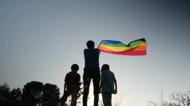 Anne ve iki çocuk gün batımında bir dağın tepesinde LGBTQ + bayrağı sallıyorlar. Aile, çeşitlilik, kapsama ve eşitlik kavramı. Gurur, lezbiyen anne ve çocuklar. Aşk aşktır.