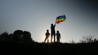 Anne ve iki çocuk gün batımında bir dağın tepesinde LGBTQ + bayrağı sallıyorlar. Aile, çeşitlilik, kapsama ve eşitlik kavramı. Gurur, lezbiyen anne ve çocuklar. Aşk aşktır.