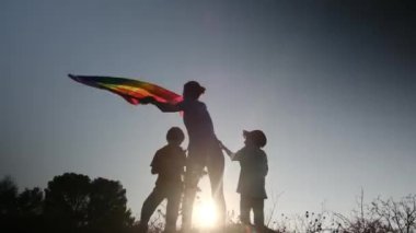 Anne ve iki çocuk gün batımında bir dağın tepesinde LGBTQ + bayrağı sallıyorlar. Aile, çeşitlilik, kapsama ve eşitlik kavramı. Gurur, lezbiyen anne ve çocuklar. Aşk aşktır.
