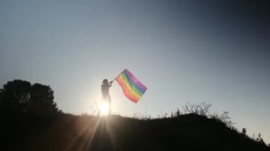 Gün batımında dalgalanan LGBTQ + bayrağıyla yalnız genç bir kadın. Eşitlik, eşitlik, dahil etme, LGBTQ konsepti. Aşk aşktır, ayrımcılık yok, özgürlüğe evet