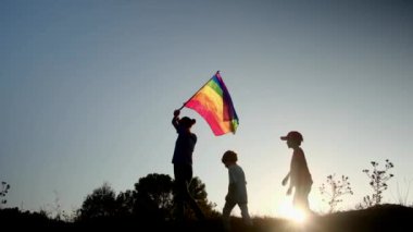 Anne ve iki çocuk gün batımında bir dağın tepesinde LGBTQ + bayrağı sallıyorlar. Aile, çeşitlilik, kapsama ve eşitlik kavramı. Gurur, lezbiyen anne ve çocuklar. Aşk aşktır.
