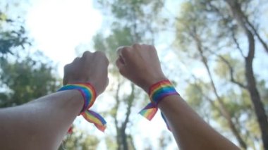 Gökkuşağı renkli LGBT bileklikle eller ve yumruklar havaya. Gurur, eşitlik, eşitlik kavramı. Güneşli bir günde gökyüzüne yumruk ve güneş ışığı yansıması. Eşitlik, kapsama, ayrımcılık yok, kabul