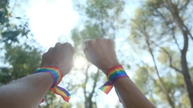Gökkuşağı renkli LGBT bileklikle eller ve yumruklar havaya. Gurur, eşitlik, eşitlik kavramı. Güneşli bir günde gökyüzüne yumruk ve güneş ışığı yansıması. Eşitlik, kapsama, ayrımcılık yok, kabul