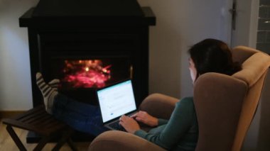Kadın şöminenin yanında dizüstü bilgisayar kullanıyor.