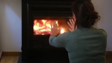 Kadın şöminenin yanında ellerini ısıtıyor.
