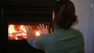 Kadın şöminenin yanında ellerini ısıtıyor.