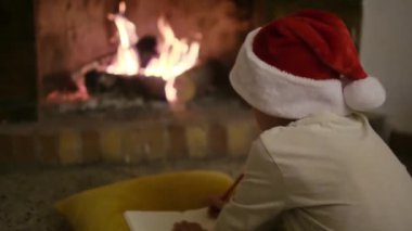 Noel Baba şapkalı çocuk şöminenin yanındaki Noel Baba 'ya mektup yazıyor.