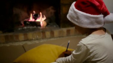 Noel Baba şapkalı çocuk şöminenin yanındaki Noel Baba 'ya mektup yazıyor.