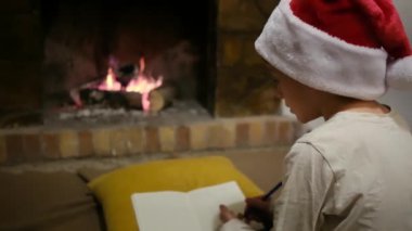 Noel Baba şapkalı çocuk şöminenin yanındaki Noel Baba 'ya mektup yazıyor.