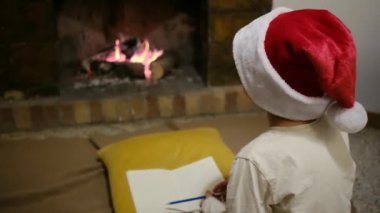 Noel Baba şapkalı çocuk şöminenin yanındaki Noel Baba 'ya mektup yazıyor.