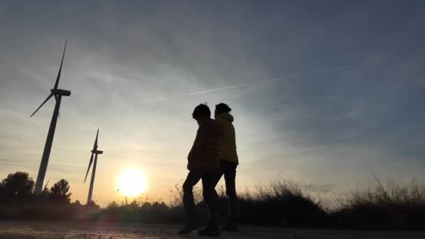 田舎で息子と一緒に日没で風力タービンに歩いている若い母親 エコロジーの概念 — ストック動画