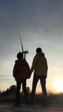 Genç anne ve oğlu kırsalda gün batımında rüzgar türbinlerine yürüyor, ekoloji kavramı.