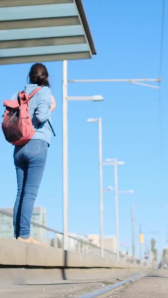 バックパックを持っている若い女性は 出発する準備ができて トラムや都市電車のプラットフォームで待っています スポーツカジュアルな服装 彼女は旅行 ルーチン 環境に優しい輸送を体現しています — ストック動画