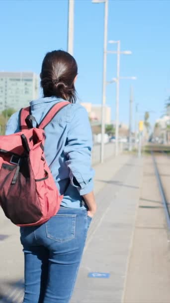 一个背着背包的年轻女子在电车或城市火车站台上等着 准备上车 她穿着休闲装 体现了旅游 日常活动 发现和环保的交通方式 — 图库视频影像