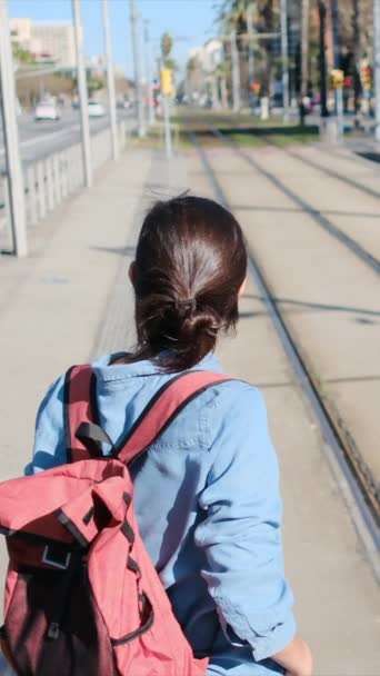 バックパックを持っている若い女性は 出発する準備ができて トラムや都市電車のプラットフォームで待っています スポーツカジュアルな服装 彼女は旅行 ルーチン 環境に優しい輸送を体現しています — ストック動画