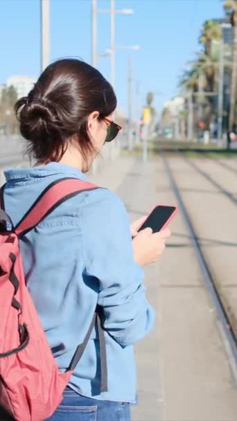 一个拿着背包的年轻女子在电车或城市火车站台上等待着 专心致志地使用手机 查看短信 电子邮件和社交媒体 她穿着休闲装 是旅游的化身 — 图库视频影像