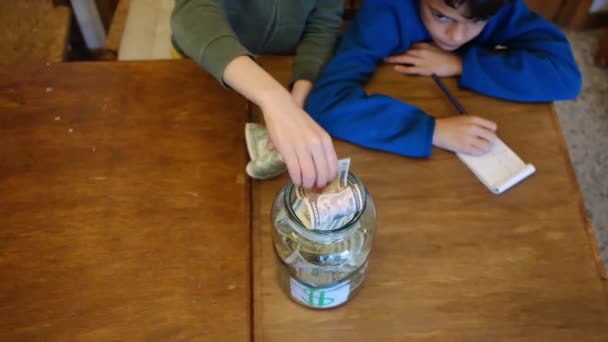 Mali Chłopcy Zbierający Banknoty Słoiku Oszczędności Siedząc Przy Drewnianym Stole — Wideo stockowe