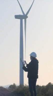 Dijital tabletli kadın elektrik mühendisi kaskını giyer güneşli bir günde rüzgar türbinini kontrol eder