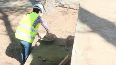 Üniforma ve kask giyen kadın işçi, güneşli bir günde inşaat alanında kaseye çimento karıştırıyor.