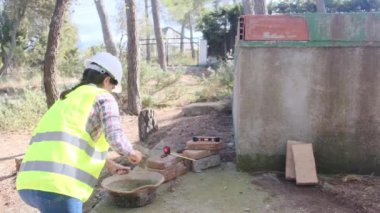Üniforma ve kask giyen kadın işçi, güneşli bir günde inşaat alanında kaseye çimento karıştırıyor.
