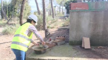 Üniforma ve kask giyen kadın işçi, güneşli bir günde inşaat alanında kaseye çimento karıştırıyor.