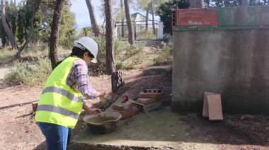 Üniforma ve kask giyen kadın işçi, güneşli bir günde inşaat alanında kaseye çimento karıştırıyor.