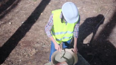 Üniforma ve kask giyen kadın işçi, güneşli bir günde inşaat alanında kaseye çimento karıştırıyor.