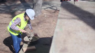 Üniforma ve kask giyen kadın işçi, güneşli bir günde inşaat alanında kaseye çimento karıştırıyor.