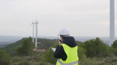 Kadın elektrik mühendisi telsizle konuşuyor ve dağlardaki rüzgar türbinlerinin yanında dijital tablet kullanıyor.