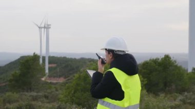 Kadın elektrik mühendisi telsizle konuşuyor ve dağlardaki rüzgar türbinlerinin yanında dijital tablet kullanıyor.