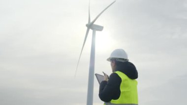 Kadın elektrik mühendisi telsizle konuşuyor ve dağlardaki rüzgar türbinlerinin yanında dijital tablet kullanıyor.