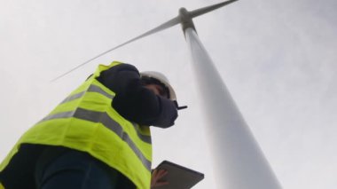 Kadın elektrik mühendisinin alt görünümü telsizle konuşuyor ve rüzgar türbini yakınında dijital tablet kullanıyor.