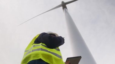 Kadın elektrik mühendisinin alt görünümü telsizle konuşuyor ve rüzgar türbini yakınında dijital tablet kullanıyor.