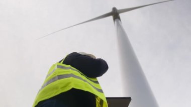 Kadın elektrik mühendisinin alt görünümü telsizle konuşuyor ve rüzgar türbini yakınında dijital tablet kullanıyor.