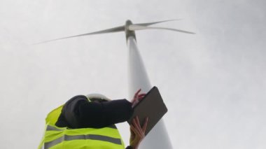 Kadın elektrik mühendisinin alt görünümü telsizle konuşuyor ve rüzgar türbini yakınında dijital tablet kullanıyor.