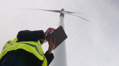 Kadın elektrik mühendisinin alt görünümü telsizle konuşuyor ve rüzgar türbini yakınında dijital tablet kullanıyor.
