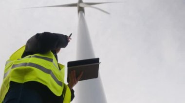 Kadın elektrik mühendisinin alt görünümü telsizle konuşuyor ve rüzgar türbini yakınında dijital tablet kullanıyor.