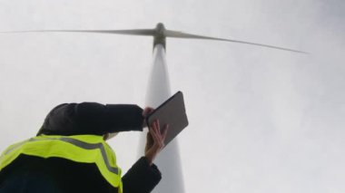 Kadın elektrik mühendisinin alt görünümü telsizle konuşuyor ve rüzgar türbini yakınında dijital tablet kullanıyor.