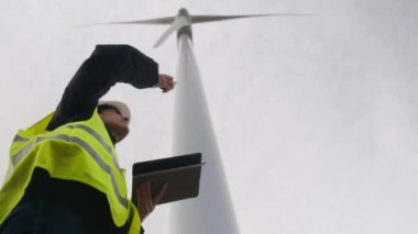 Kadın elektrik mühendisinin alt görünümü telsizle konuşuyor ve rüzgar türbini yakınında dijital tablet kullanıyor.