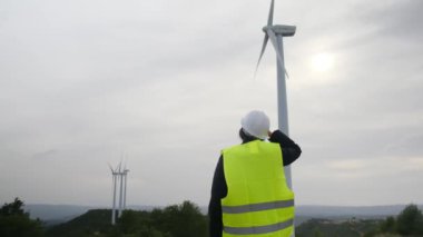 Üniformalı ve kasklı kadın elektrik mühendisinin dağlardaki rüzgar türbinlerine bakışı.