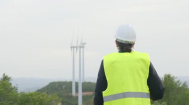 Üniformalı ve kasklı kadın elektrik mühendisinin dağlardaki rüzgar türbinlerine bakışı.
