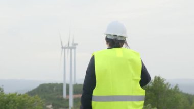 Üniformalı ve kasklı kadın elektrik mühendisinin dağlardaki rüzgar türbinlerine bakışı.
