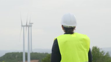 Üniformalı ve kasklı kadın elektrik mühendisinin dağlardaki rüzgar türbinlerine bakışı.