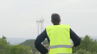 Üniformalı ve kasklı kadın elektrik mühendisinin dağlardaki rüzgar türbinlerine bakışı.